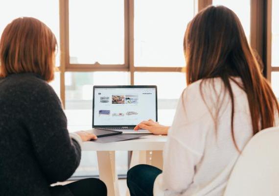 Two persons look at content displayed in a laptop. Their back is to the camera.